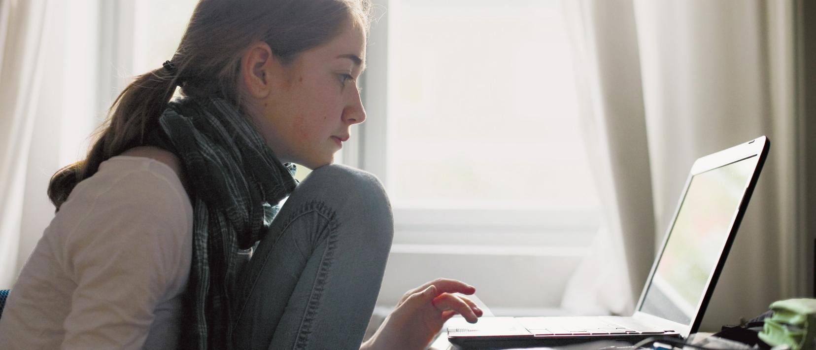 Eine junge Frau sitzt konzentriert und mit angezogenen Beinen vor ihrem Laptop und tippt etwas ein.