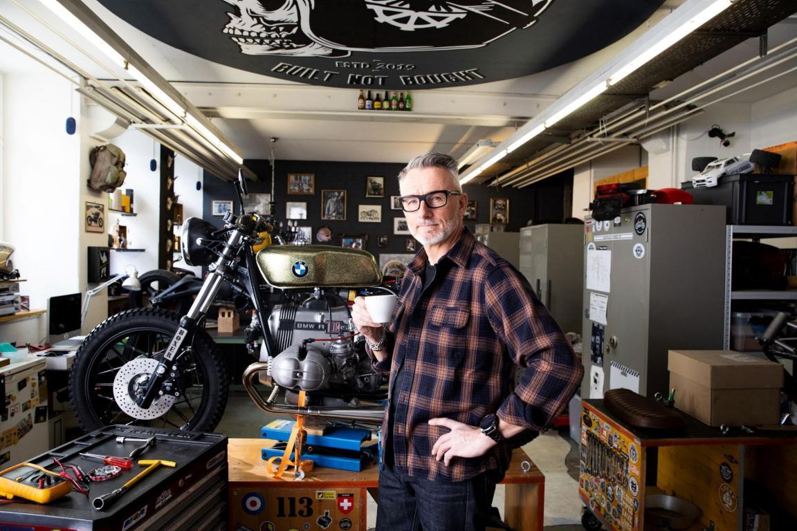 Mitarbeiter in der Freizeit mit Kaffee in der Hand in der Motorradwerkstatt mit Motorrad im Hintergrund auf der Hebebühne und Werkzeuge davor liegend.