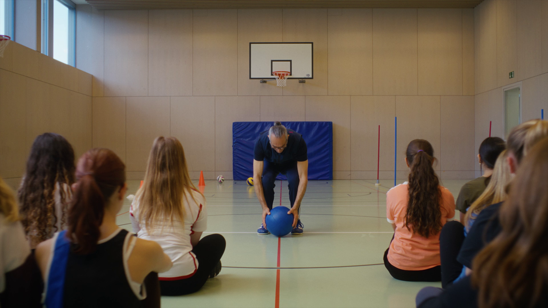 Sportlehrer in einer Sporthalle zeigt der Schulklasse eine Übung mit einem Medizinball vor.