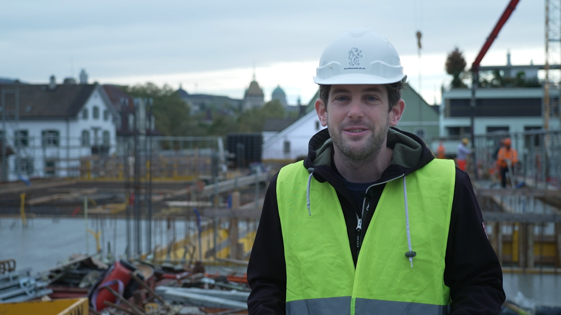Unser Mitarbeiter steht mit einem weissen Helm auf dem Kopf sowie einer gelben Sicherheitsweste auf einer Baustelle. Im Hintergrund sind Armierungseisen zu sehen sowie Baufachleute, welche Beton giessen. Weiter im Hintergrund sind zahlreiche städtische Gebäude zu sehen.
