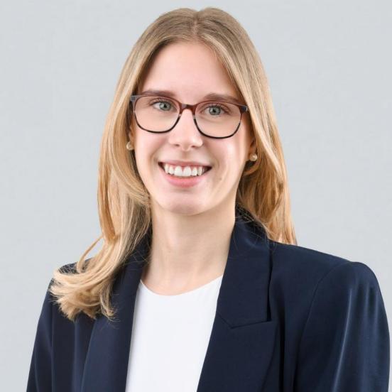 Ein Portraitbild, das unsere Mitarbeiterin mit langen blonden Haaren, einer dunkeln Brille, Perlenohrstecker sowie einem dunkeln Blazer zeigt.