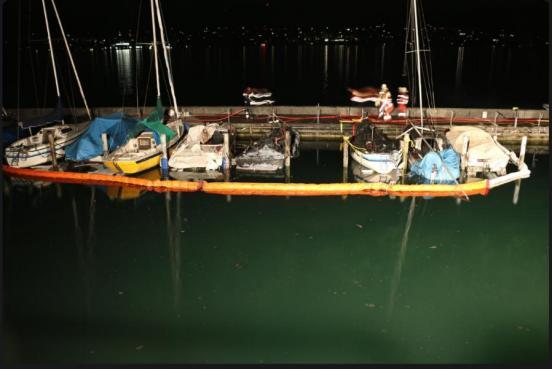 Sieben in Brand geratene Boote nebeneinander an einem Steg vertaut und mit einer Ölsperre um ringt.