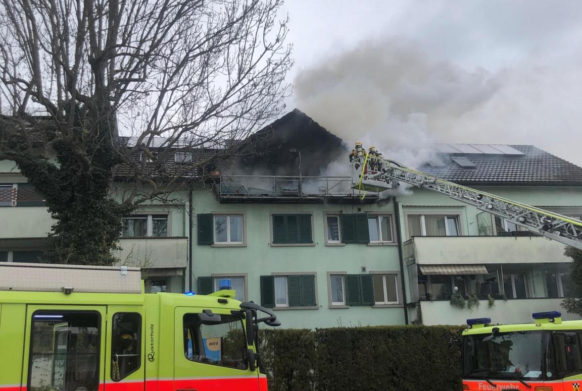 Dichter Rauch aus Dachwohnung eines Mehrfamilienhauses und Löschkräfte bei der Arbeit