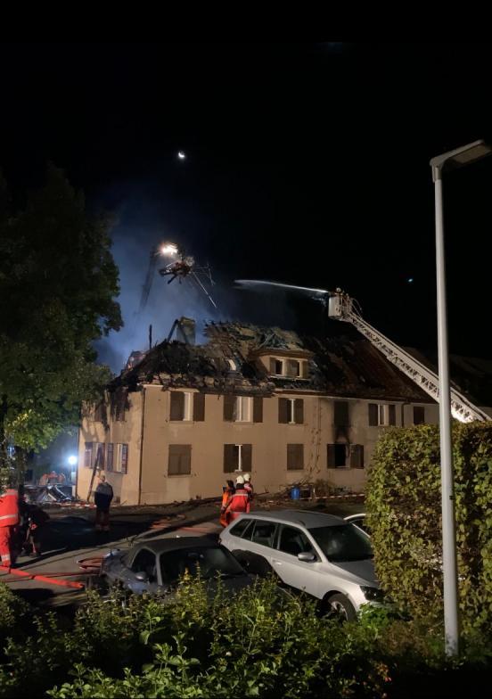 Mehrfamilienhaus mit ausgebranntem Dachstock und der Feuerwehr an den Löscharbeiten