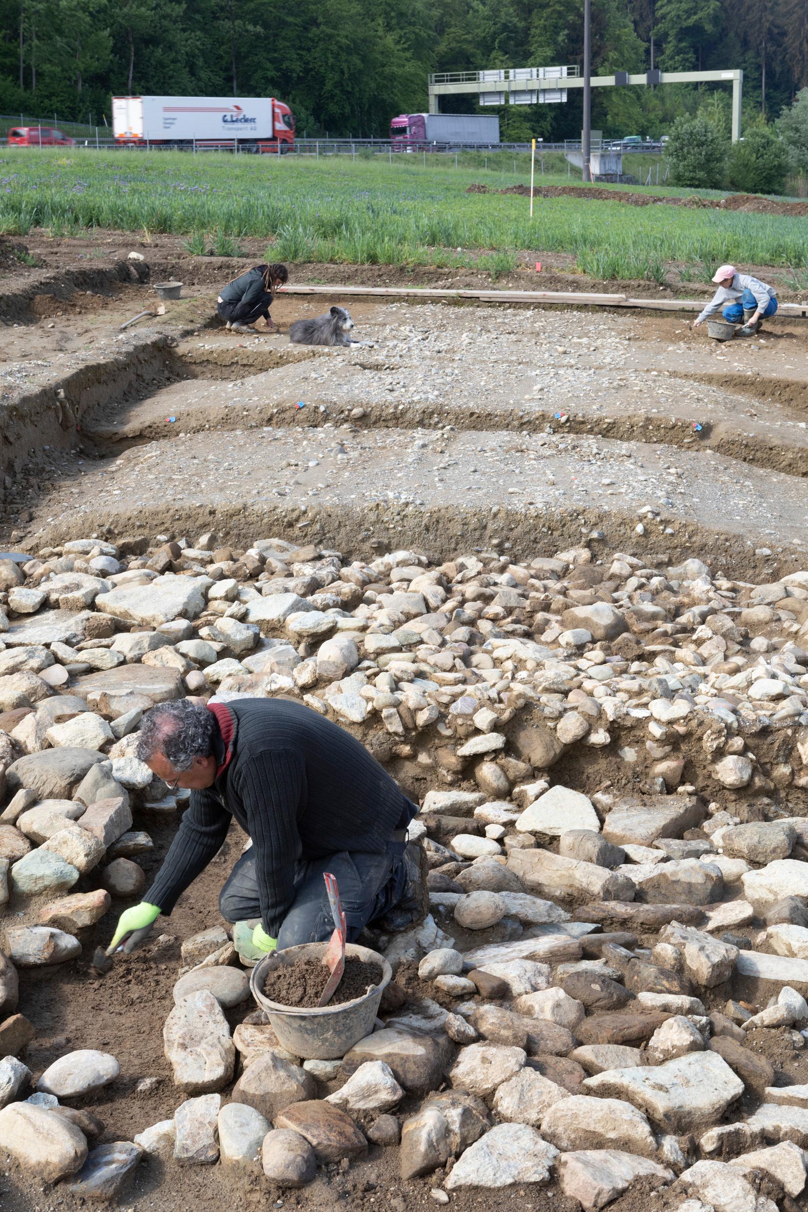 Ausgräber und Archäologinnen legen die römische Fernstrasse bei Wiesendangen frei. Im Hintergrund rauscht ihre moderne Nachfolgerin, die Autobahn A1. 