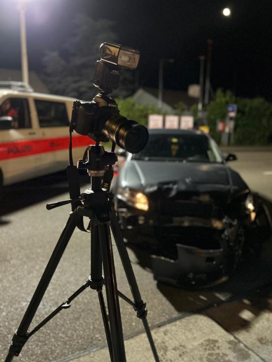 Fotokamera auf Stativ im Vordergrund mit Unfallfahrzeug und Polizeiauto im Hintergrund