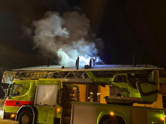Ein Feuerwehrauto steht vor dem brennenden Haus; auf dem Dach bekämpften Feuerwehrmänner den Brand