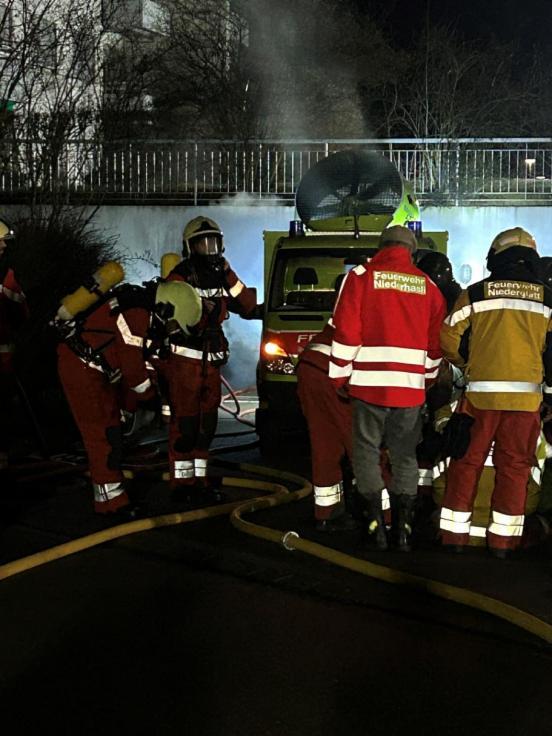 Feuerwehrleute mit einem Grosslüfter im Hintergrund vor einer Tiefgarageneinfahrt, aus der Rauch kommt.