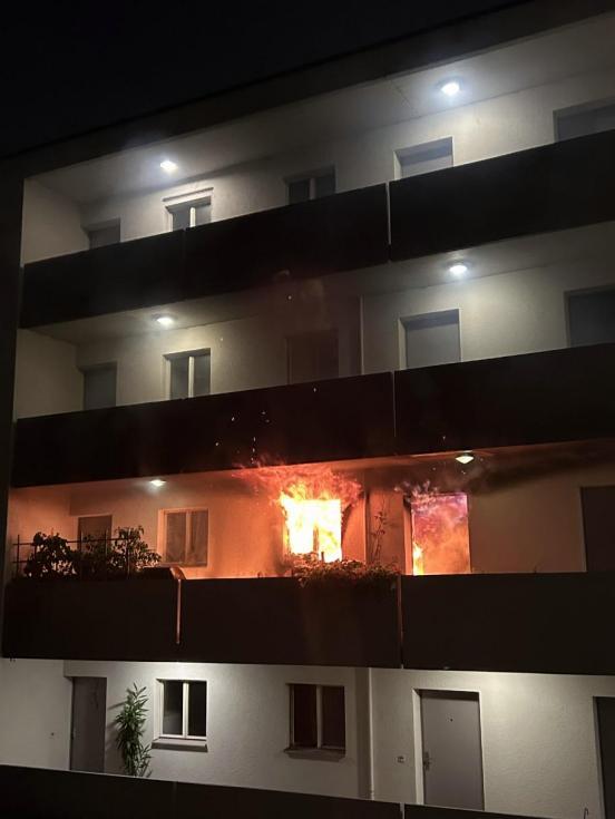 Feuer dringt aus einer Wohnung eines Mehrfamilienhauses. Es ist dunkle Nacht. 