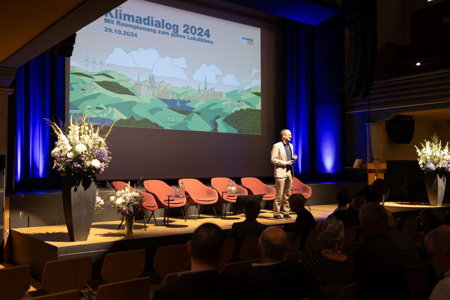 Martin Neukom auf der Bühne, im Hintergrund eine Illustration zum Klimadialog.