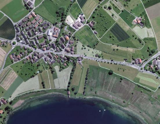 Blick von oben auf ein kleines Dorf an einem See; durch das Dorf gibt es eine wellenlinig-geführte Strasse