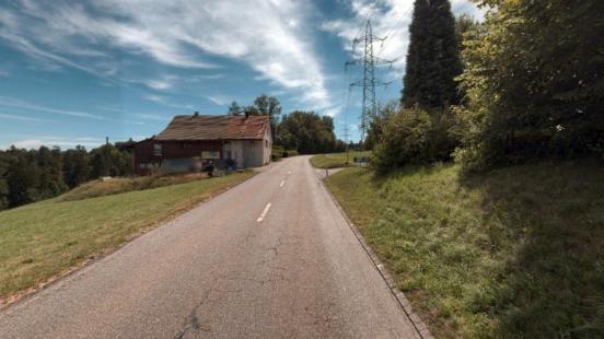 Bergaufführende Strasse mit Wiese und einem Bauernhaus auf der linken Seite, rechts ist eine Böschung mit Bäumen und Sträuchern und eine Hochspannungsleitung.