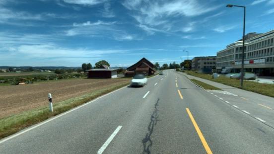 Strasse mit drei Spuren, die durch eine weisse und eine gelbe gestrichelte Linie getrennt sind und links durch eine Wiese so-wie rechts durch einen Radweg und Industriegebäude begrenzt wird.
