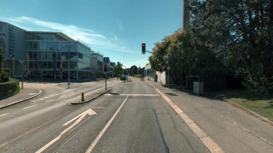 Eine Strassenkreuzung mit Markierungen und einem Lichtsig-nal; eine Strasse zeigt nach links ab. Im Hintergrund steht ein Bürogebäude mit einer Glasfront
