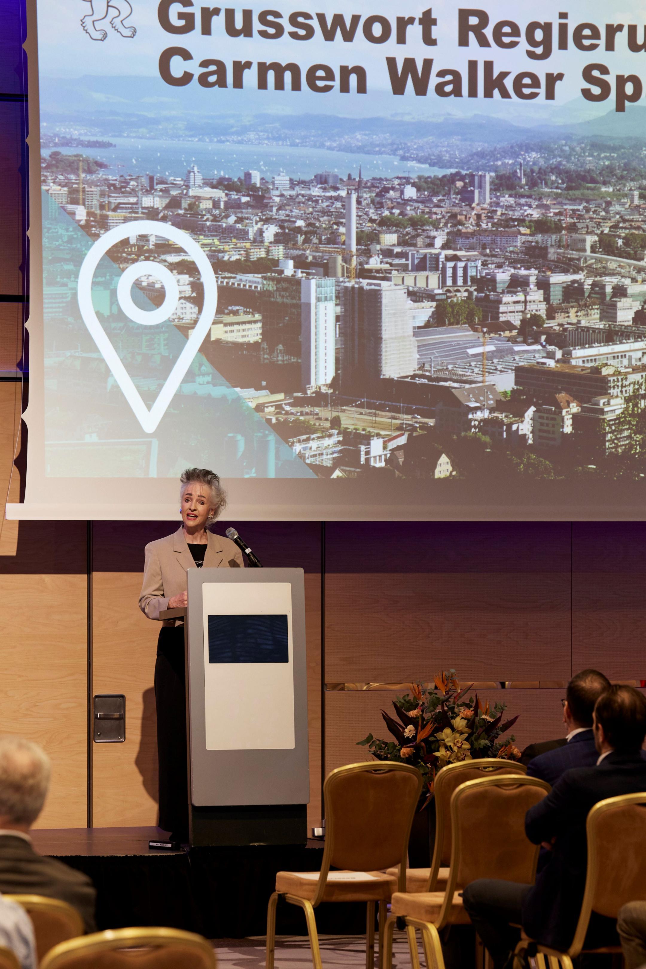Regierungsrätin Carmen Walker Späh steht auf einer Bühne hinter einem Podest und spricht ins Publikum. Auf der Leinwand ist eine Luftaufnahme der Stadt Zürich zu sehen.
