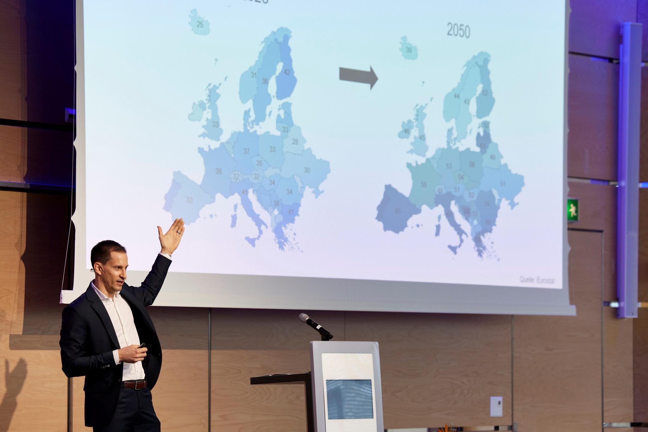 Der Amtsleiter des Amts für Wirtschaft trägt einen schwarzen Anzug, steht auf einer Bühne und deutet auf eine Leinwand hinter ihm. Dort sind Zahlen zu sehen, wie sich das Verhältnis von Menschen im erwerbsfähigen Alter zu Menschen im nichterwerbsfähigen Alter in verschiedenen Ländern in Europa bis ins Jahr 2050 entwickelt.