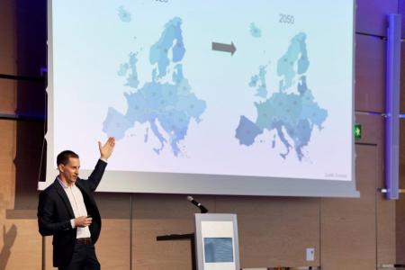 Der Amtsleiter des Amts für Wirtschaft trägt einen schwarzen Anzug, steht auf einer Bühne und deutet auf eine Leinwand hinter ihm. Dort sind Zahlen zu sehen, wie sich das Verhältnis von Menschen im erwerbsfähigen Alter zu Menschen im nichterwerbsfähigen Alter in verschiedenen Ländern in Europa bis ins Jahr 2050 entwickelt.