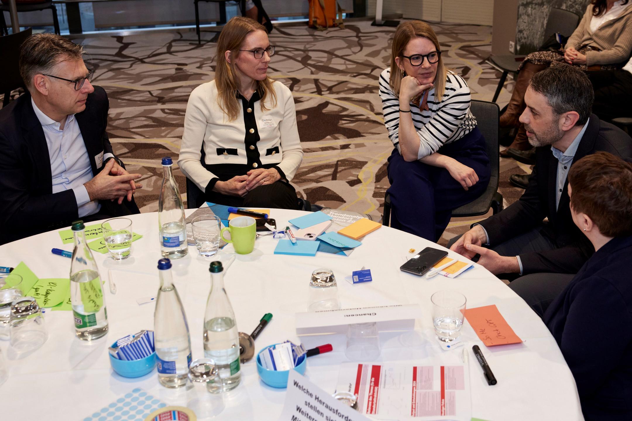 Fünf Personen sitzen an einem runden Tisch, auf dem farbige Post-it liegen. Sie diskutieren angeregt miteinander..