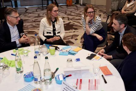 Fünf Personen sitzen an einem runden Tisch, auf dem farbige Post-it liegen. Sie diskutieren angeregt miteinander..