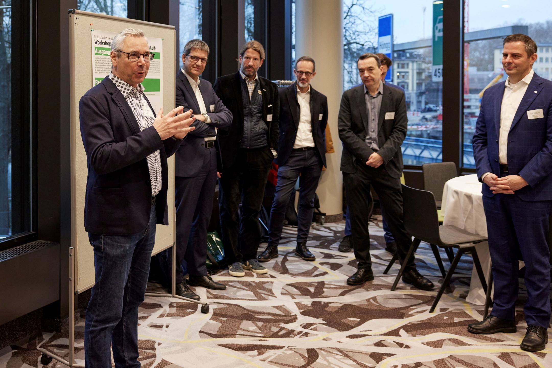 Sechs Männer in Anzügen stehen im Kreis und sprechen miteinander.