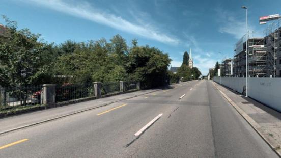 Gerade Strasse mit einer ge-strichelten weissen Mittellinie und einer gelben gestrichelten Linie auf der linken Seite. Links ist ein Zaun mit Bäumen dahin-ter und rechts ist eine Baustelle für neue Wohnblöcke.