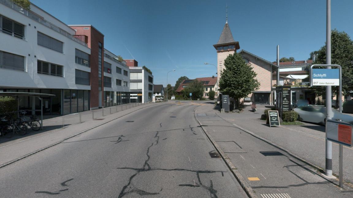 Strasse mit schwarzen Flecken, links ein weisser Wohnblock mit einem überdachten Veloständer vorne dran, rechts ein Trottoir und eine Tafel für eine Bushaltestelle.