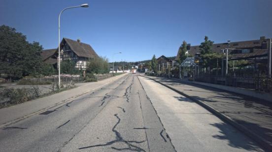 Ein Strasse mit einer Laterne und einem Riegelhaus auf der linken Seite; rechts ist eine Bushaltestelle.