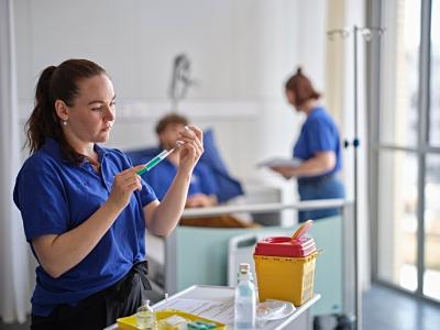 Pflegefachkraft in blauer Uniform bereitet eine Injektion vor, im Hintergrund befinden sich zwei weitere Pflegepersonen in einem hellen Patientenzimmer.