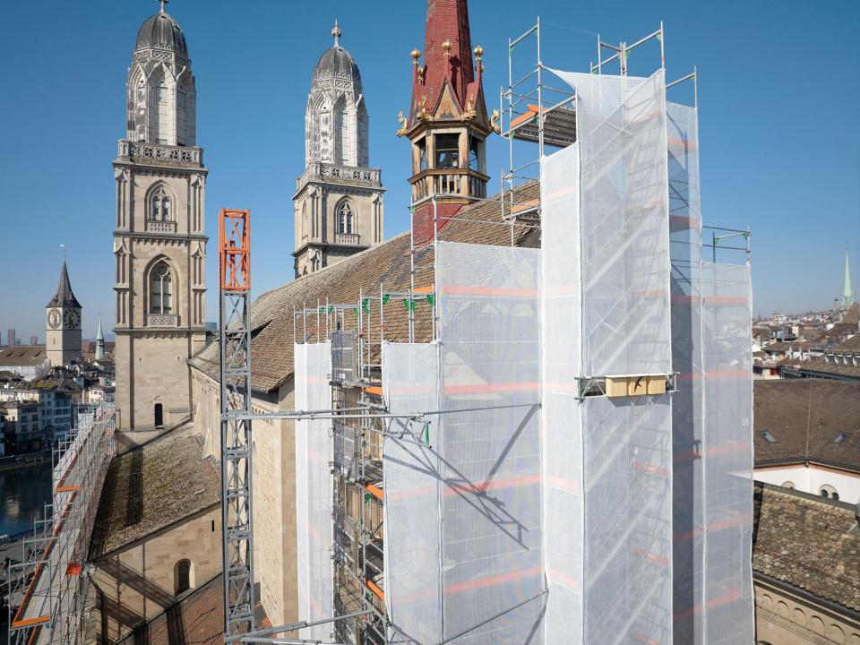 Gerüst mit weissem Netz umgeben. Die markanten Doppeltürme des romanischen Bauwerks ragen links und in der Mitte des Bildes auf. Im Hintergrund ist ein Blick auf die Stadt Zürich mit weiteren Kirchtürmen und Gebäuden unter einem klaren, blauen Himmel zu sehen.