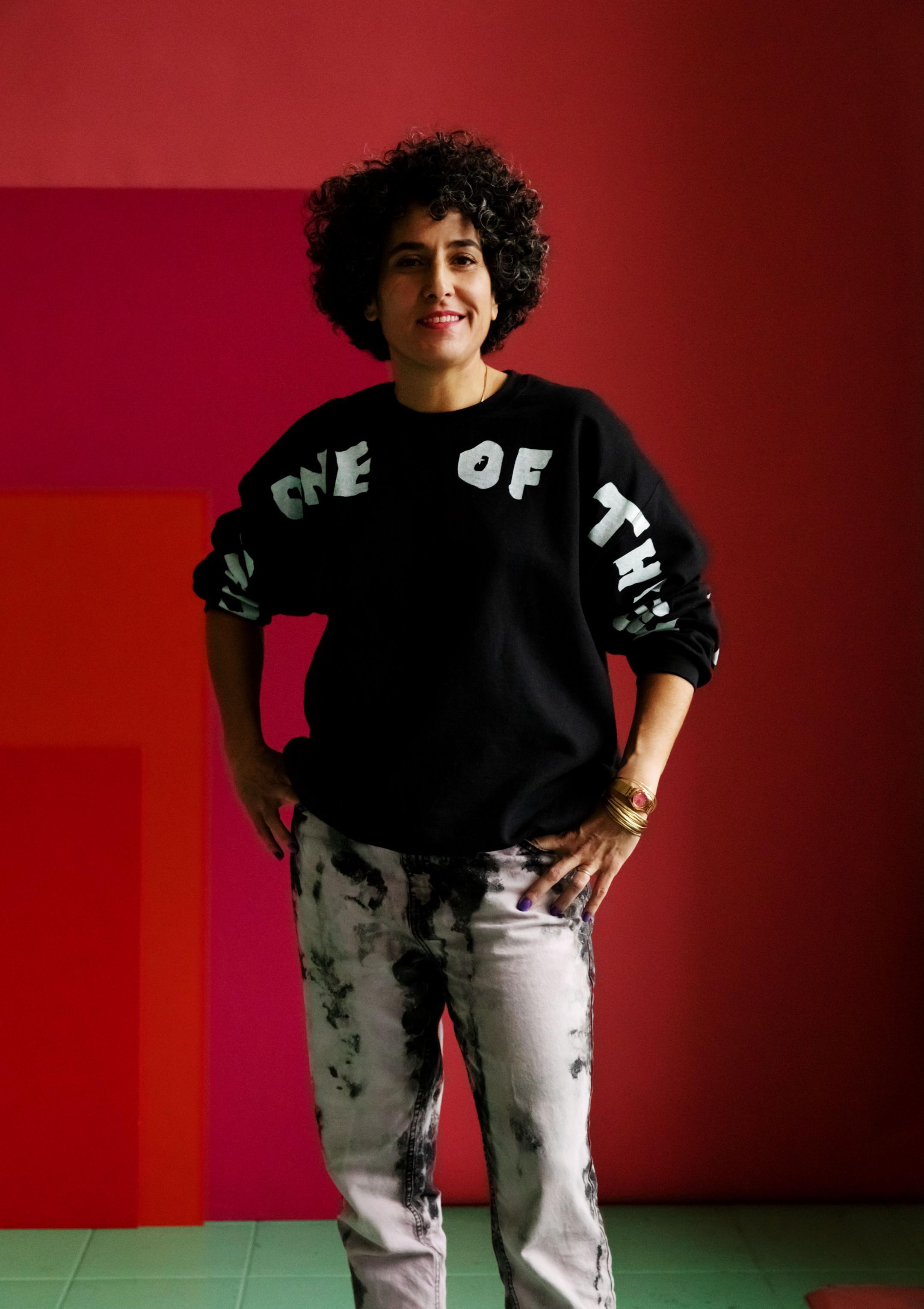 Eine Frau mit lockigem, dunklem Haar und einem schwarzen Pullover mit weißer Aufschrift posiert 