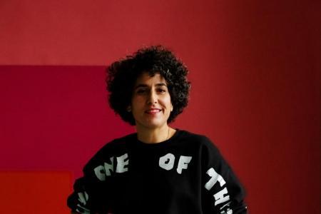 Eine Frau mit lockigem, dunklem Haar und einem schwarzen Pullover mit weißer Aufschrift posiert 