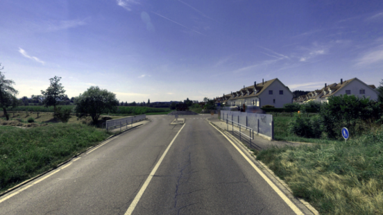 Asphaltierte Landstrasse mit durchgezogener Mittellinie, die auf eine Kreuzung mit einem Kreisel zuführt. Wohnhäuser mit Giebeldächern auf der rechten Seite, Felder und Vegetation auf der linken Seite. Blauer Himmel mit vereinzelten Wolken.