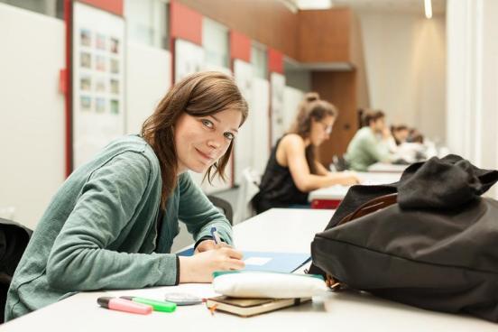 Portraitaufnahme einer Schülerin im Klassenzimmer. Die junge Frau sitzt, vor ihr auf dem Pult liegen ihre Lernunterlagen und Stifte.