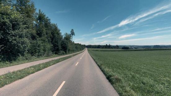 Blick auf eine asphaltierte Strasse mit Mittelstreifen, die sich durch eine ländliche Gegend schlängelt. Links der Strasse dichter Wald, rechts ein weites grünes Feld. Der Himmel ist blau mit vereinzelten weissen Wolken.