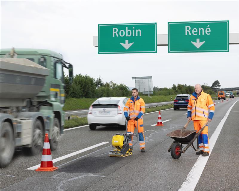 Auf einer mehrspurigen Autobahn arbeiten zwei Männer in orangefarbener Arbeitskleidung an einer beschädigten Stelle. Einer bedient eine Rüttelplatte, der andere schiebt eine Schubkarre. Über ihnen hängen grüne Wegweiser mit den Namen «Robin» und «René» und Pfeilen nach unten. Im Hintergrund fahren Autos und ein LKW.
