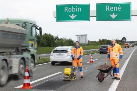 Auf einer mehrspurigen Autobahn arbeiten zwei Männer in orangefarbener Arbeitskleidung an einer beschädigten Stelle. Einer bedient eine Rüttelplatte, der andere schiebt eine Schubkarre. Über ihnen hängen grüne Wegweiser mit den Namen «Robin» und «René» und Pfeilen nach unten. Im Hintergrund fahren Autos und ein LKW.