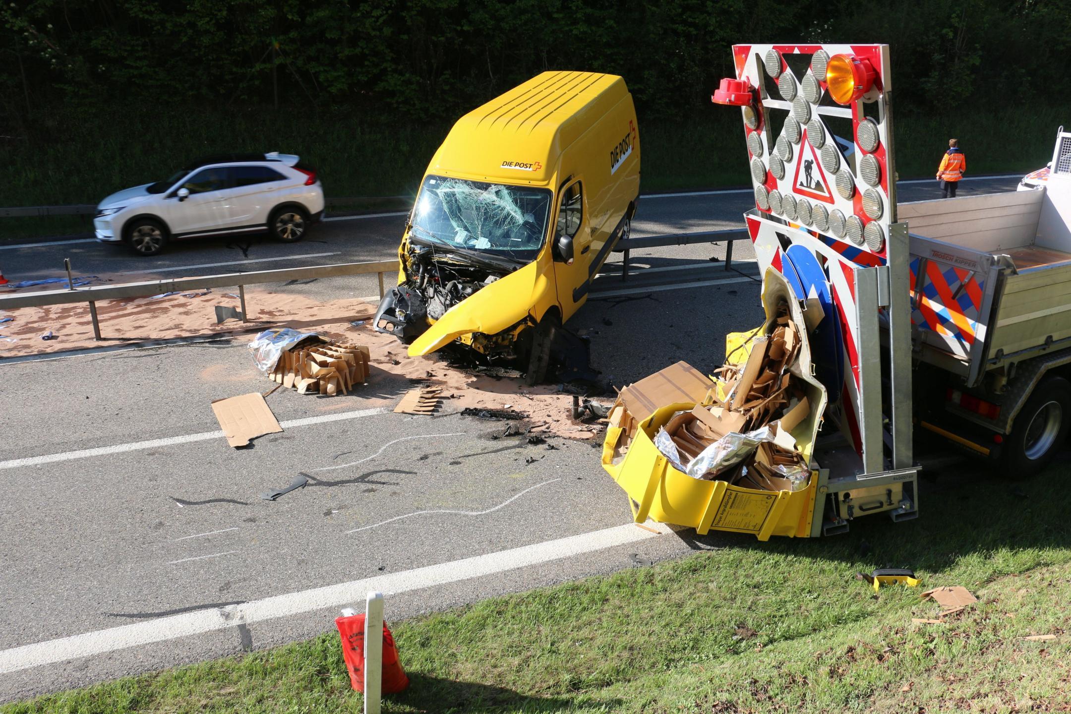 Ein schwer beschädigter gelber Lieferwagen steht quer zur Fahrbahn auf einer Autobahn. Trümmer und Kartons sind verstreut. Ein Lastwagen mit einer Warntafel steht daneben. Im Hintergrund ein weißes Auto und eine Person in orangefarbener Kleidung.