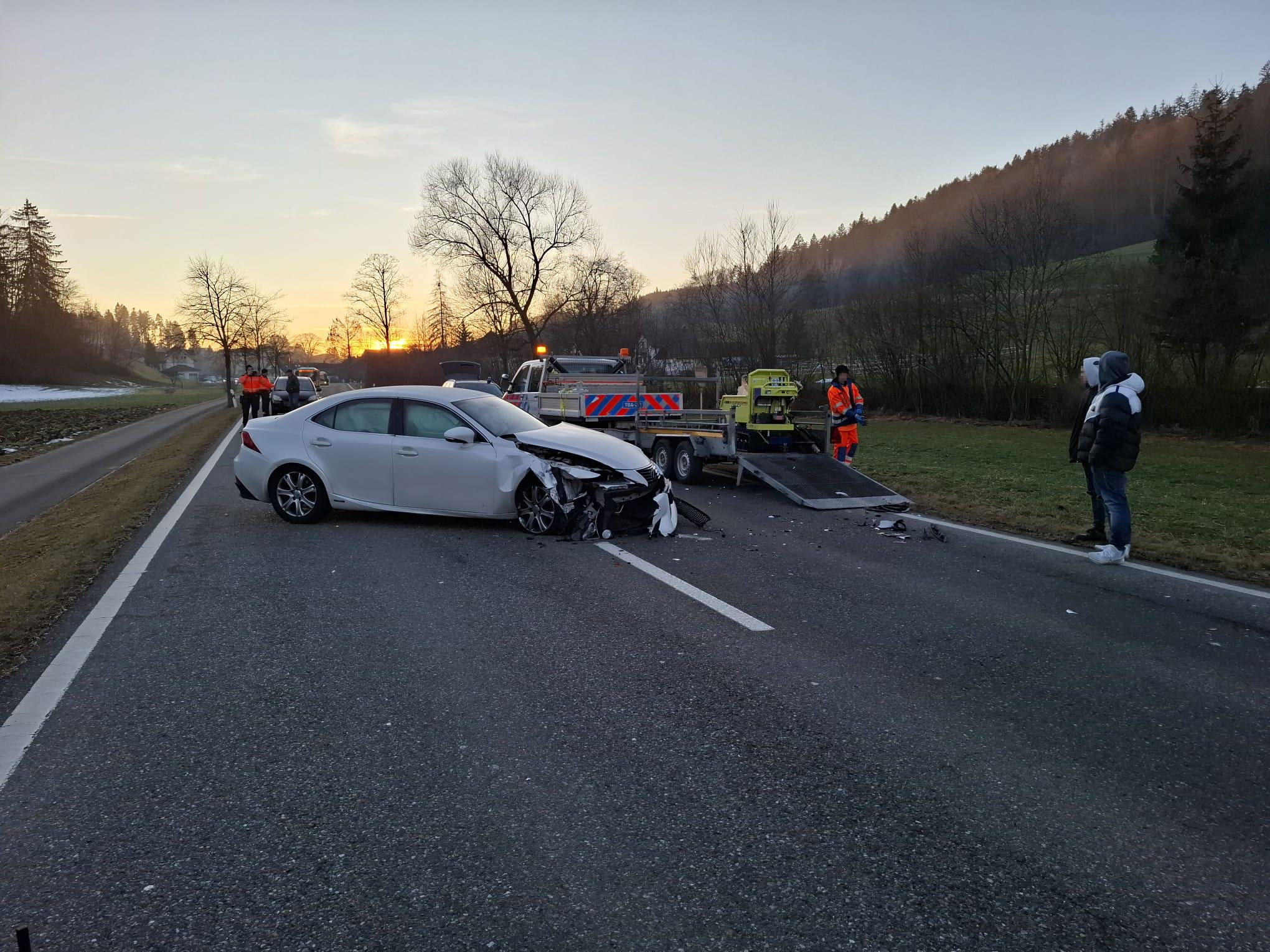 Ein weisser Personenwagen mit erheblichen Frontschäden steht nach einem Unfall quer auf einer Landstrasse. Ein Abschleppwagen ist vor Ort, und mehrere Personen, möglicherweise Einsatzkräfte, befinden sich in der Nähe.