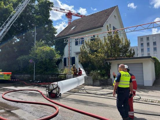 Ein mobiler Lüchtungsschlauch der Feuerwehr führt in ein Einfamilienhaus. Davor stehen ein Polizist und ein Feuerwehrmann