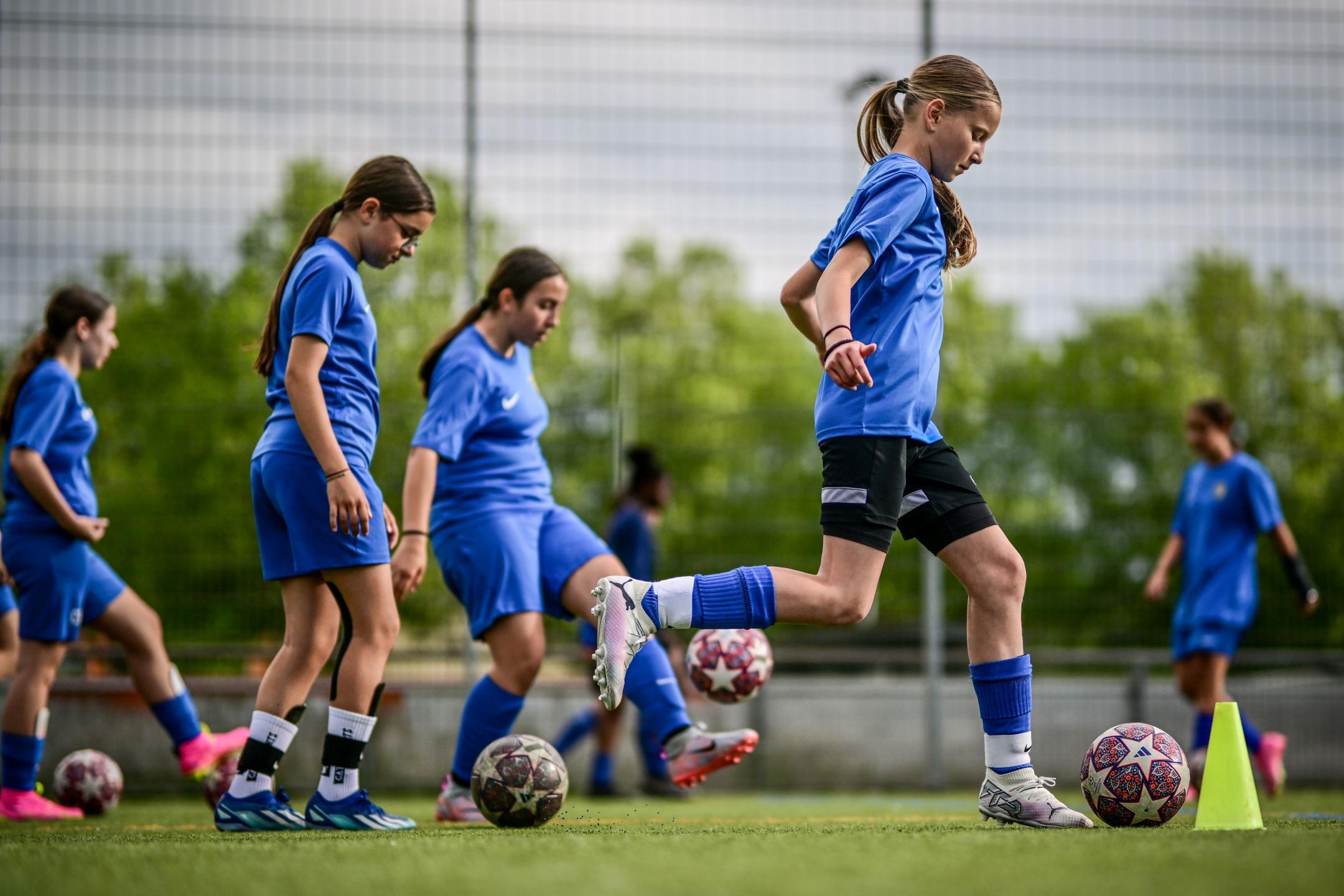 Sechs Mädchen in blauen Fussballtrikots trennen mit dem Fussball am Fuss über einen Kunstrasenplatz.