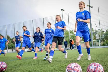 Sieben Mädchen in blauen Fussballtrikots rennen in einer Reihe ohne Ball über einen Kunstrasenplatz.
