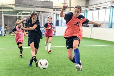 Vier Mädchen in Fussball-Trainingskleidung rennen einem Fussball auf Kunstrasen in einer Halle nach.