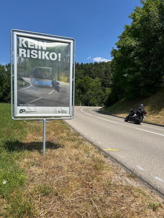 Eine Tafel mit einem Motorrad-Kampagnenplakat, auf welchem «Kein Risiko» steht., neben einer Strasse, auf welcher ein Motorrad in Kurvenlage fährt.