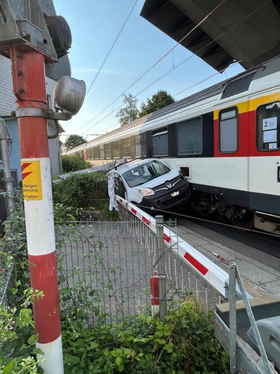 Auto ist zwischen Zug und einer Gartenmauer eingeklemmt, davor befindet sich eine geschlossene Bahnschranke