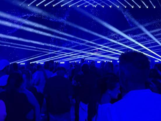 Menschen tanzen in einem blauen Raum. An der Decke sind Scheinwerferstrahlen zu sehen.