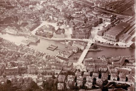 Ein alter, schwarz-weisser Luftbildblick auf eine europäische Stadt. Ein grosser Fluss oder See teilt die Stadt in mehrere Teile, die durch Brucken verbunden sind. 