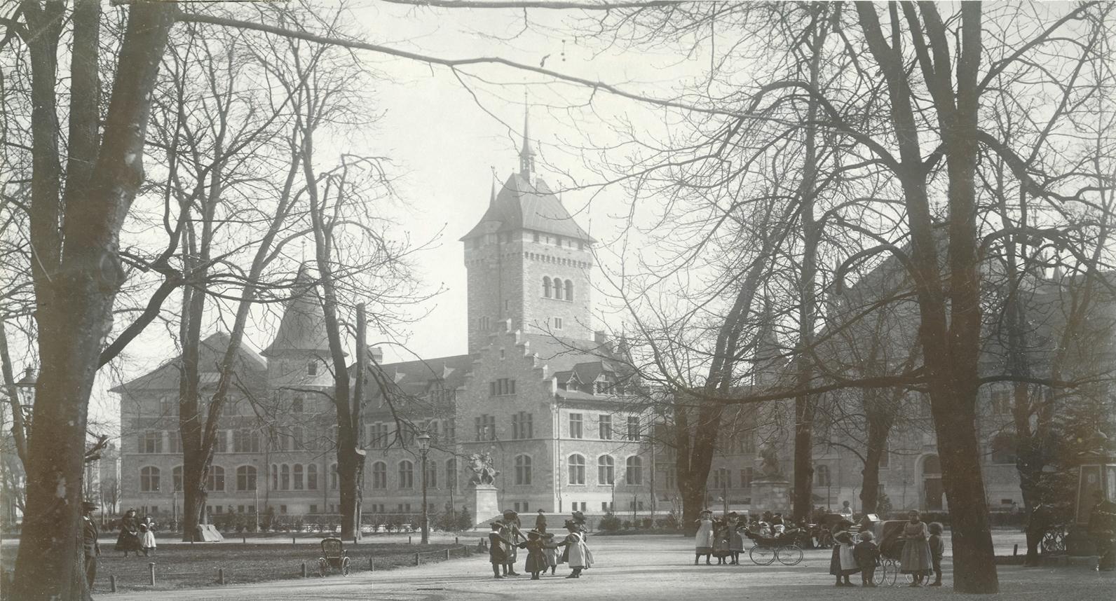 Ein altes Schwarz-Weiss-Foto, das ein grosses, historisches Gebaude mit einem markanten Turm zeigt. Es ist von kahlen Baumen umgeben.