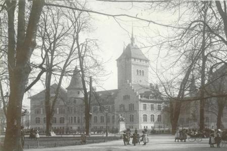 Ein altes Schwarz-Weiss-Foto, das ein grosses, historisches Gebaude mit einem markanten Turm zeigt. Es ist von kahlen Baumen umgeben.