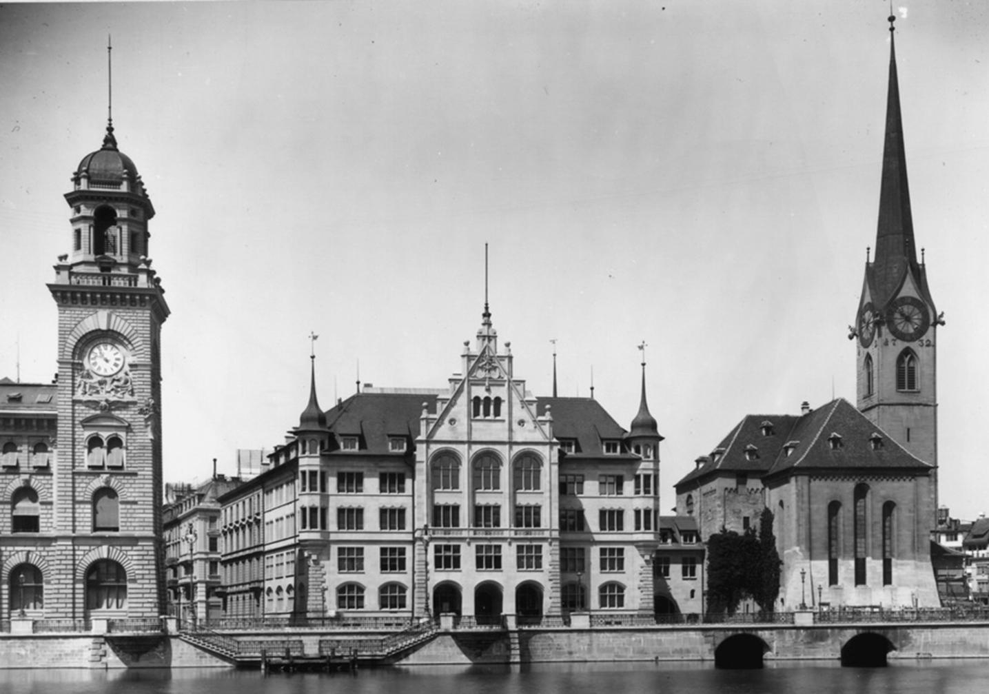 Ein altes, Schwarz-Weiss-Foto einer beeindruckenden Gebäudereihe an einem Flussufer. Auffallend sind zwei markante Türme: links ein grosser Uhrturm mit einer Kuppel und rechts ein schlanker Kirchturm mit einem Spitzdach.