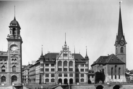 Ein altes, Schwarz-Weiss-Foto einer beeindruckenden Gebäudereihe an einem Flussufer. Auffallend sind zwei markante Türme: links ein grosser Uhrturm mit einer Kuppel und rechts ein schlanker Kirchturm mit einem Spitzdach.